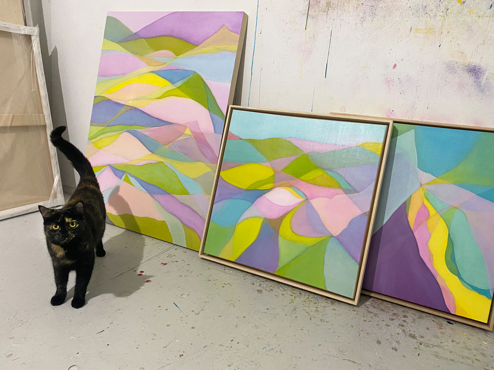 dark tortoiseshell cat stands beside 3 abstract paintings leaning against paint splattered studio wall.